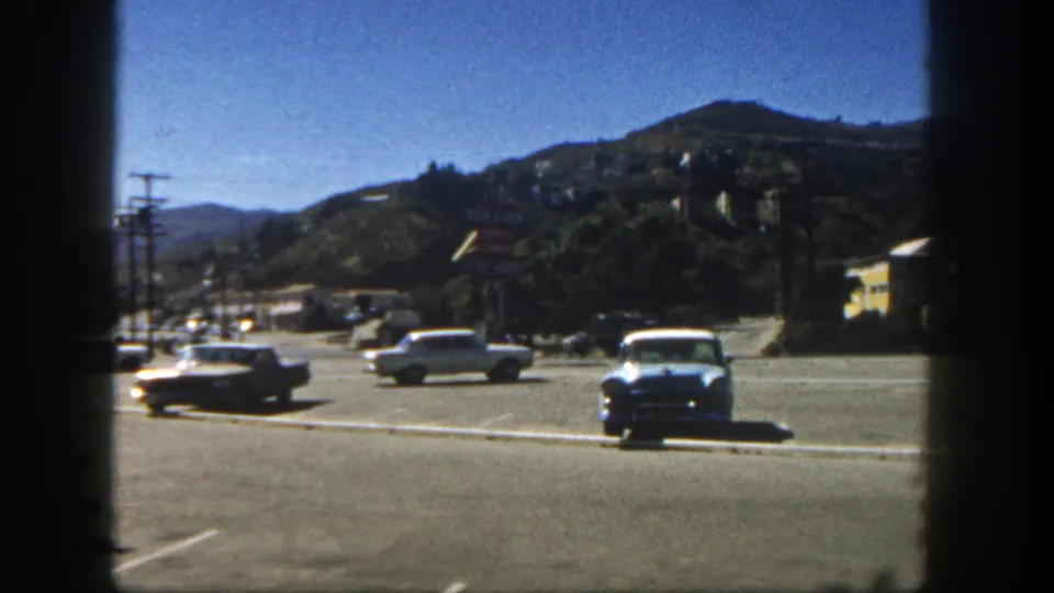 1962 San Diego parking lot with a blue sedan, passing cars, painted lines, and hillside houses in the background.