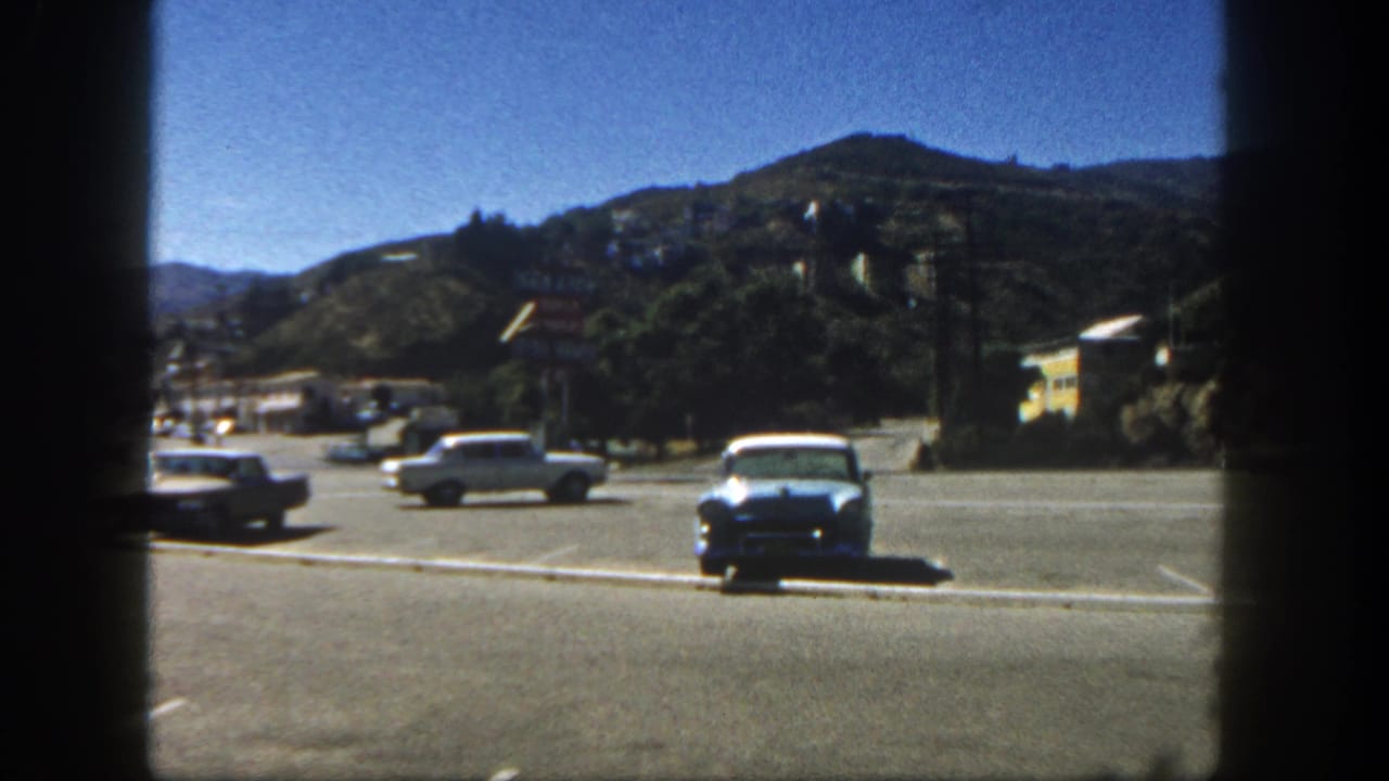 Cars cross a paved lot with hillside houses in the distance.