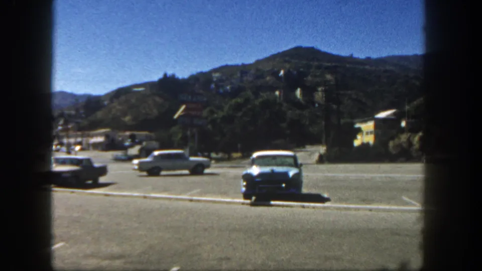 1962 San Diego view of classic cars in a paved parking area framed by dark film edges and a hillside backdrop.
