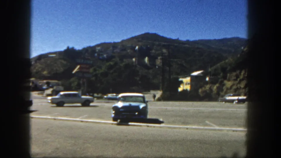 1962 San Diego parking-lot frame with a blue sedan, moving cars, painted spaces, and a residential hillside beyond.