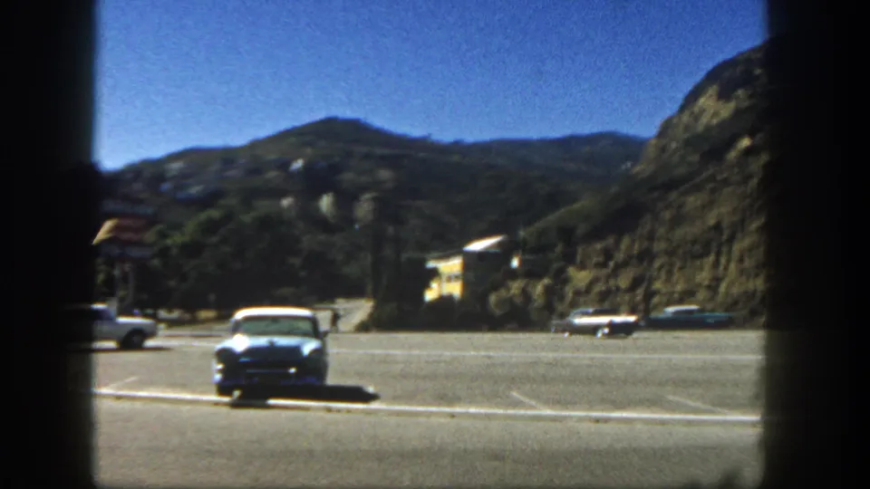 1962 San Diego parking-lot view with a blue sedan, distant cars, hillside houses, and dark vertical framing at the edge.