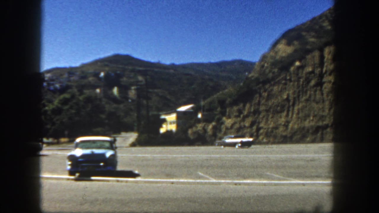 1962 San Diego home-movie frame showing a roadside area near a hillside with a white car, mountains, and bright sky.