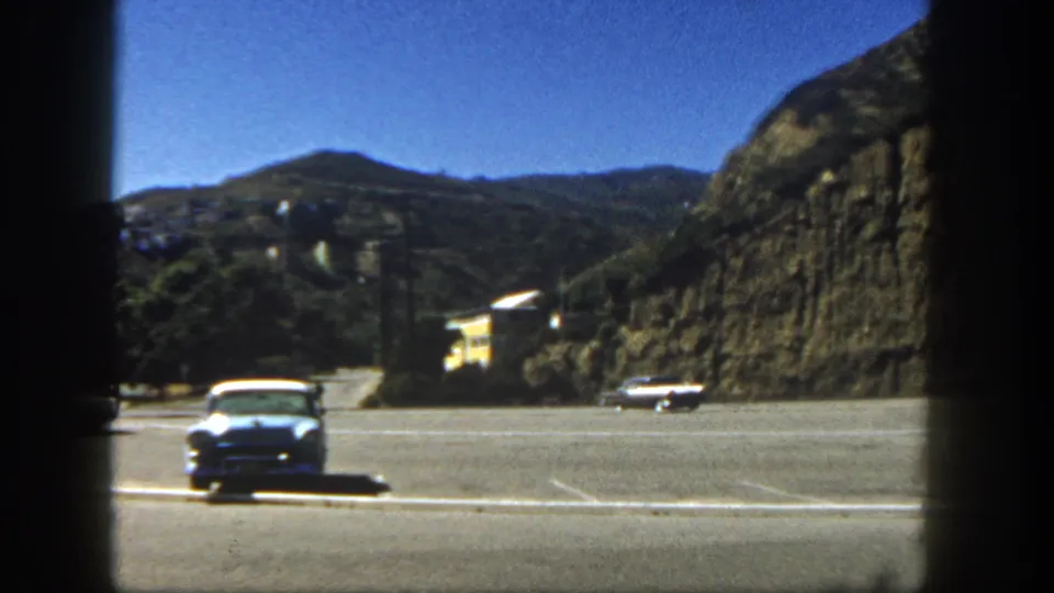 1962 San Diego home-movie frame showing a roadside area near a hillside with a white car, mountains, and bright sky.