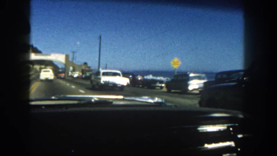 1962 San Diego road scene from inside a car, showing coastal traffic, parked vehicles, road signs, and ocean water.