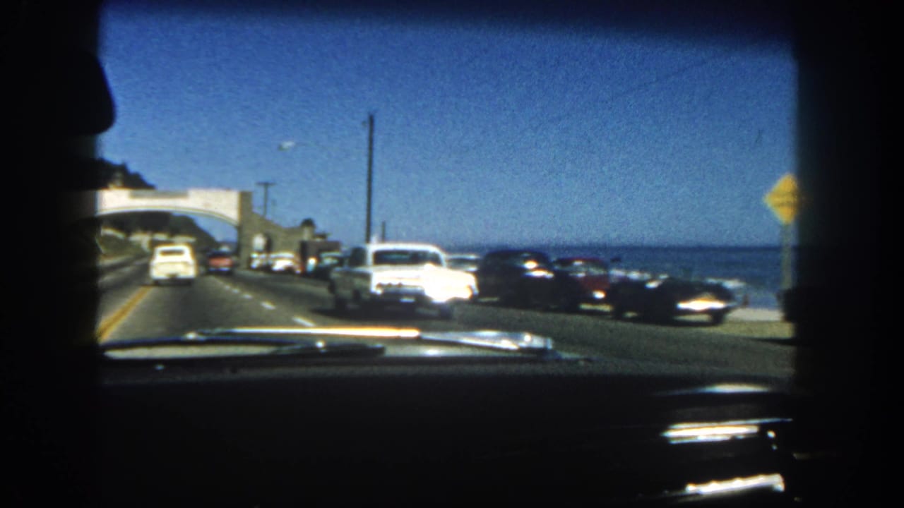 1962 San Diego coastal highway view from inside a car, with classic traffic ahead, ocean to the right, dashboard trim, and a dark passenger profile.