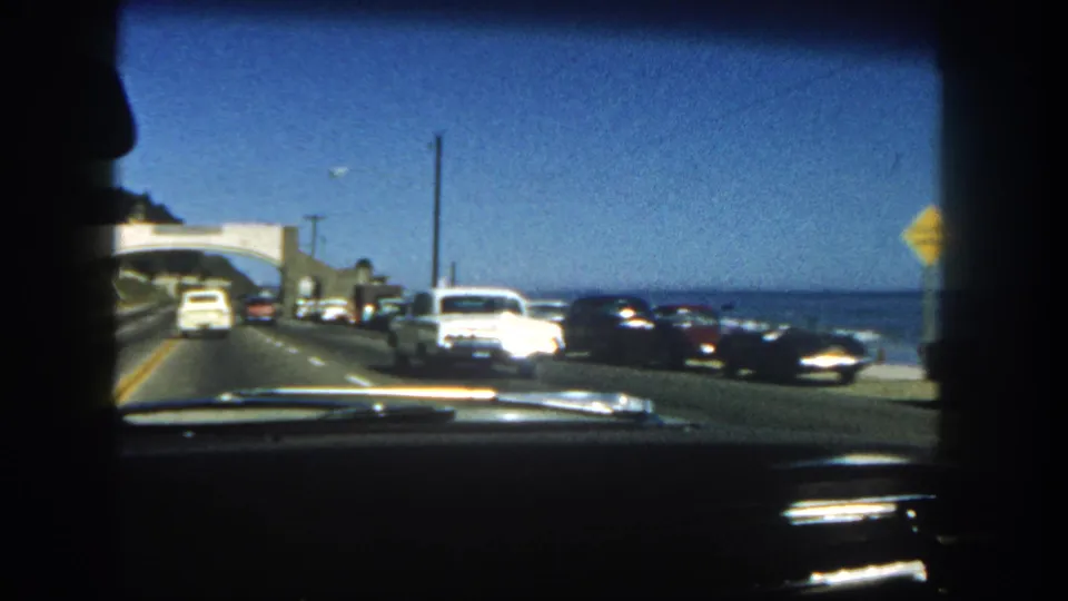 1962 San Diego coastal highway view from inside a car, with classic traffic ahead, ocean to the right, dashboard trim, and a dark passenger profile.