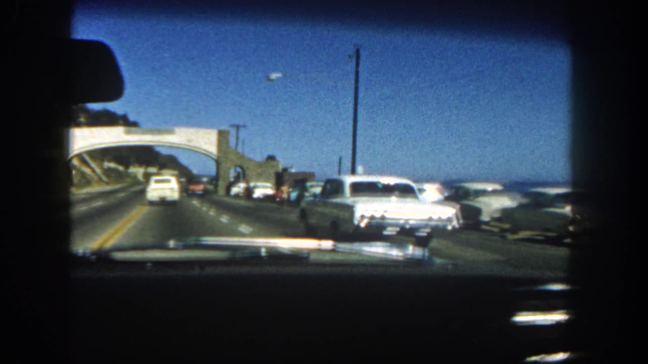 Inside-car view of a 1962 San Diego coastal drive, with a white sedan passing near the camera, traffic ahead, ocean at right, and an overpass ahead.