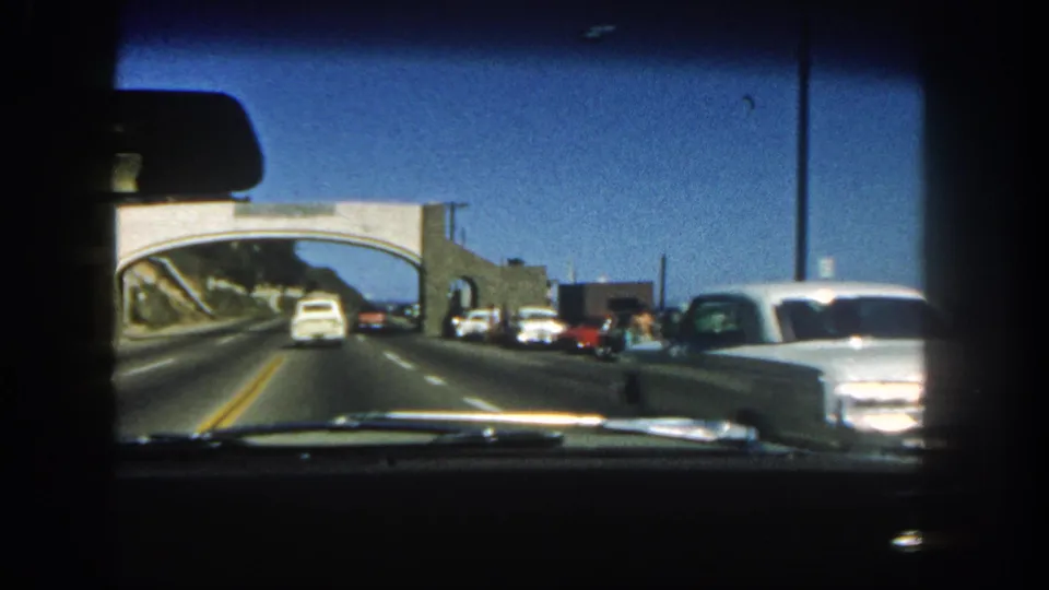 1962 San Diego road footage from a car interior, showing traffic ahead, a concrete overpass, roadside buildings, and a white sedan along the right side.
