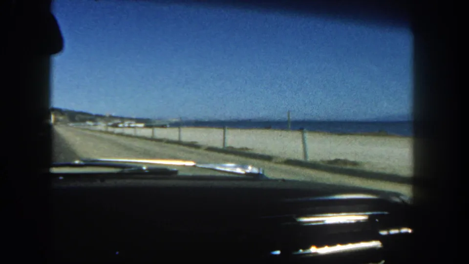 Final 1962 San Diego coastal highway view from inside the car, with open pavement, low roadside barrier, blue ocean, dashboard, and passenger silhouette.