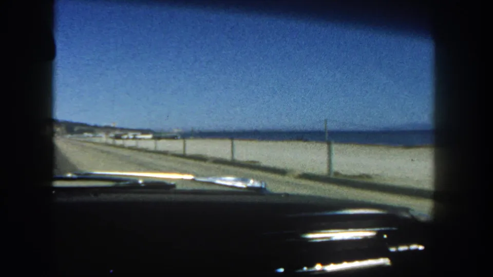 1962 San Diego 8mm frame from inside a car, looking down a beachside road with ocean, low wall, dashboard, and blue sky visible.