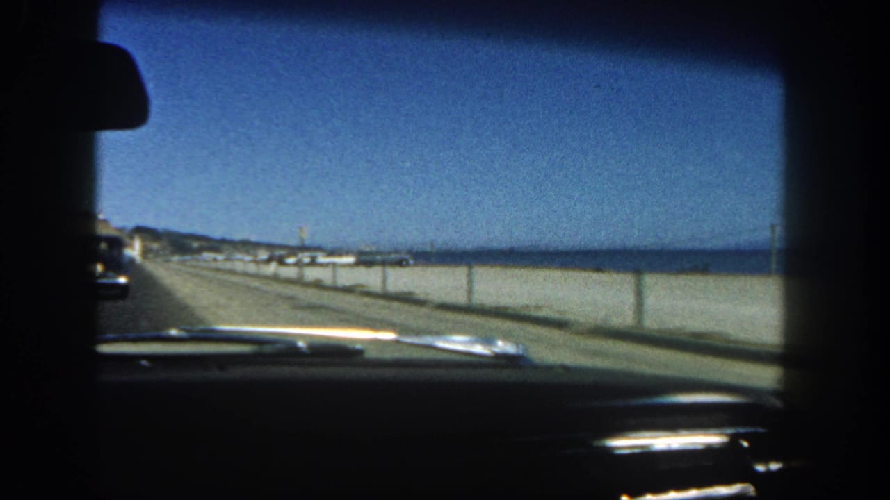 Car interior foreground with a beach road, ocean, and another vehicle ahead.