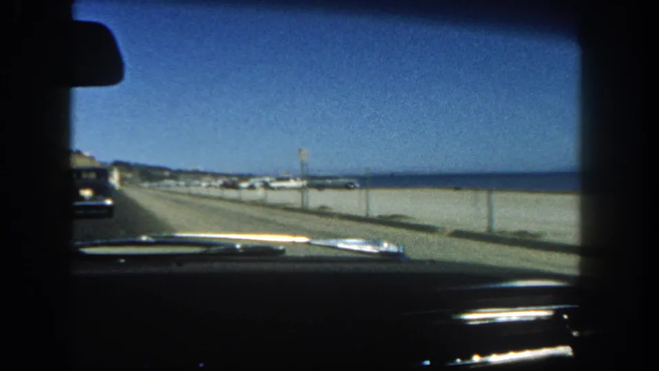 1962 San Diego coastal drive frame with dashboard foreground, beach road, low wall, ocean, distant shoreline buildings, and another car at left.