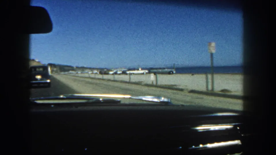 Inside a 1962 car in San Diego, the windshield frames a coastal road, dark vehicle ahead, roadside sign, beach, ocean, and distant buildings.