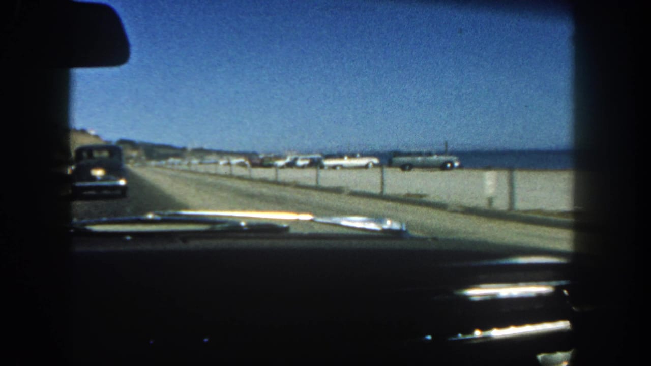 View through a car windshield along a coastal road with parked cars and ocean beyond.