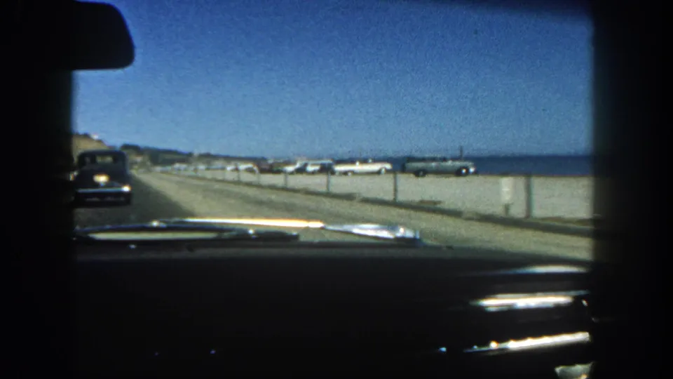 1962 San Diego-area home-movie view from inside a car, looking past the dashboard toward a beach road, parked cars, and ocean horizon.