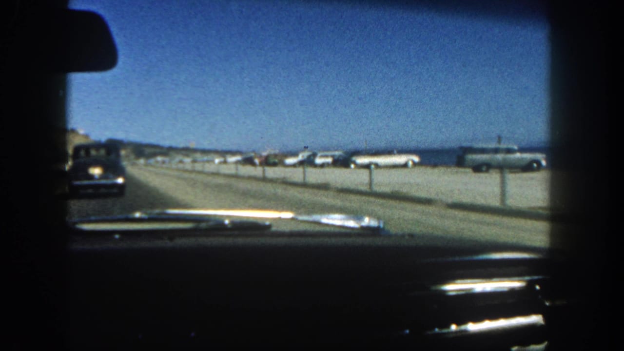 A windshield view on a San Diego coastal road in 1962 shows parked classic cars, a low beach barrier, and blue water in the distance.