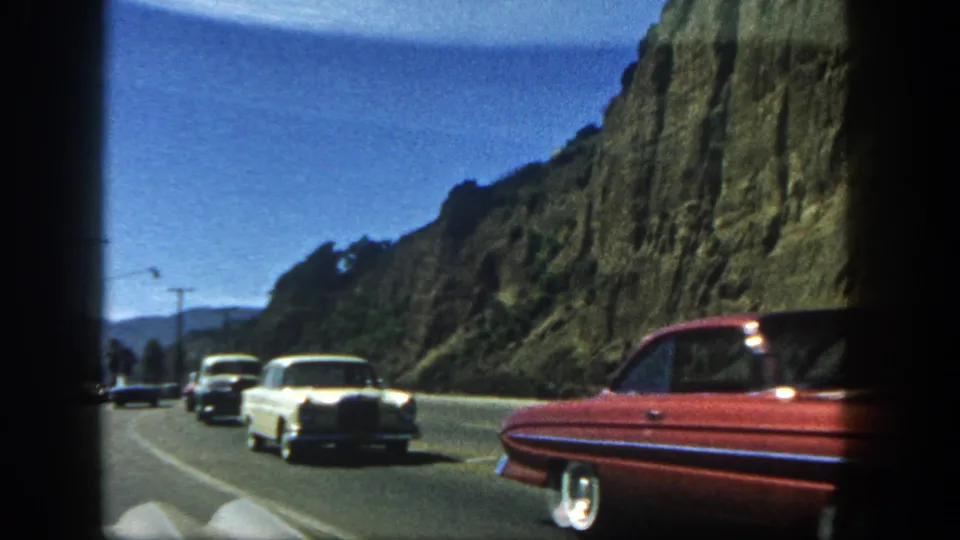 1962 home-movie frame of a winding Southern California road beside a rugged cliff, with mid-century cars moving through the curve.