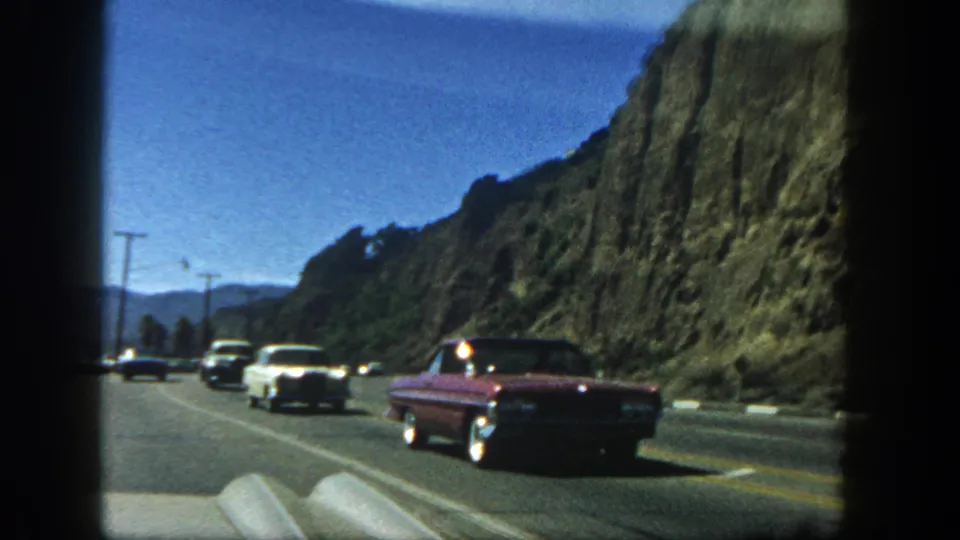 A red mid-century car passes on a cliffside road in 1962 San Diego-area footage, with other vehicles and rugged hills in view.