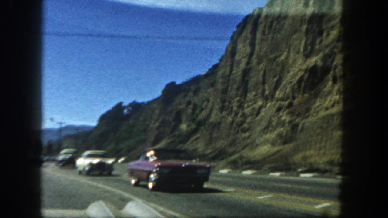 1962 San Diego home-movie view of classic cars on a multilane highway next to a rugged cliff, seen through film frame edges.