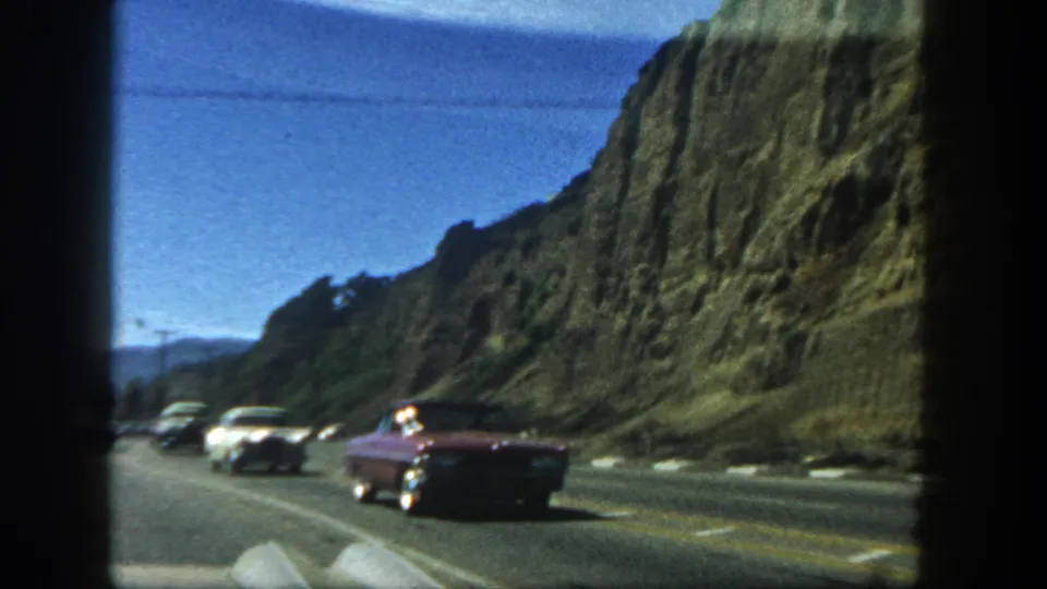 1962 San Diego home-movie view of classic cars on a multilane highway next to a rugged cliff, seen through film frame edges.