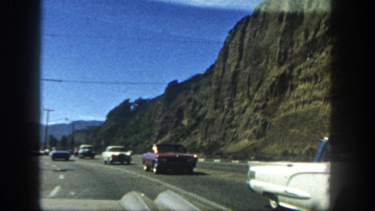 Passenger-view 1962 San Diego highway footage showing mid-century cars, a rocky hillside, utility lines, and a partial white car at the edge.