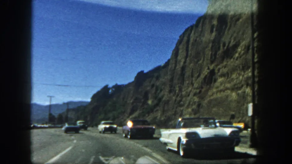 1962 San Diego road scene with a white convertible, classic cars, utility poles, and a cliffside highway filmed from a moving vehicle.