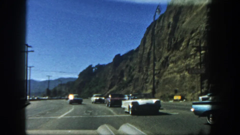 Late clip frame from 1962 San Diego showing classic cars on a broad highway beside a rocky hillside, with saturated color and visible film edges.