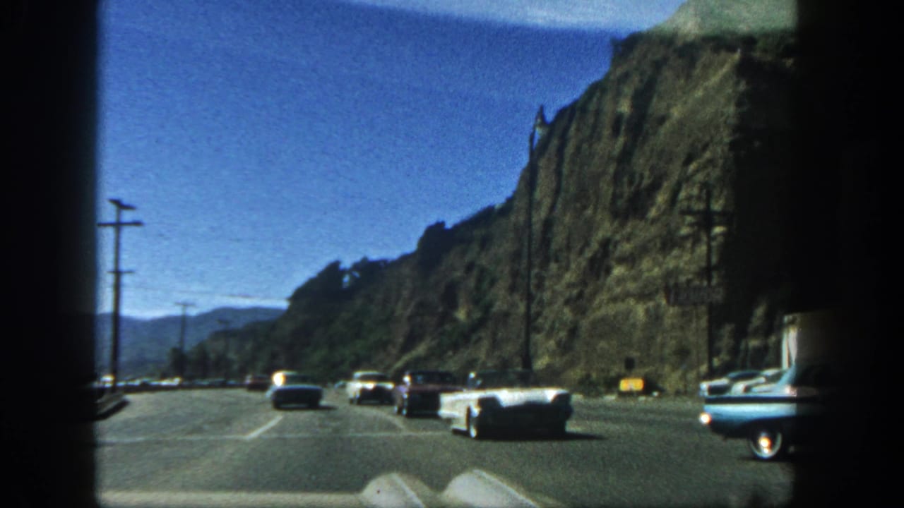 Classic cars drive along a paved road beside a steep rocky cliff.