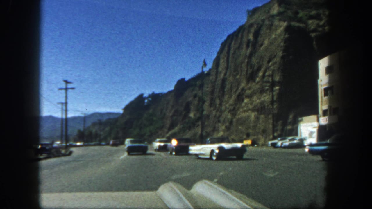 1962 San Diego driving view with a white convertible and other cars moving along a rocky roadside.
