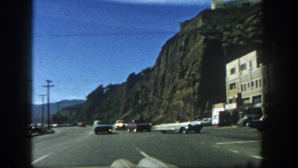 1962 San Diego home-movie frame showing classic cars, a roadside building, utility poles, and a steep rock face.