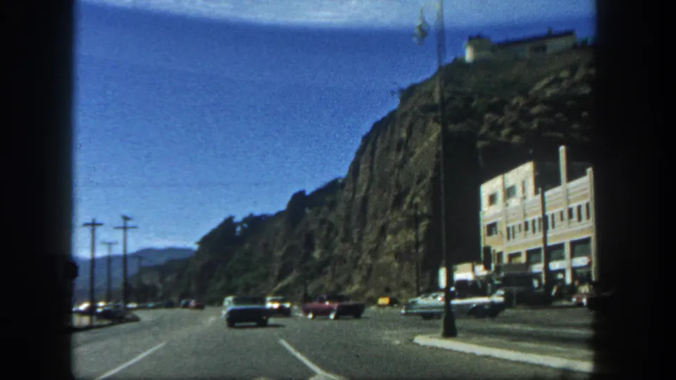 1962 San Diego driving footage frame with classic cars, a cliffside building, utility poles, and distant hills.
