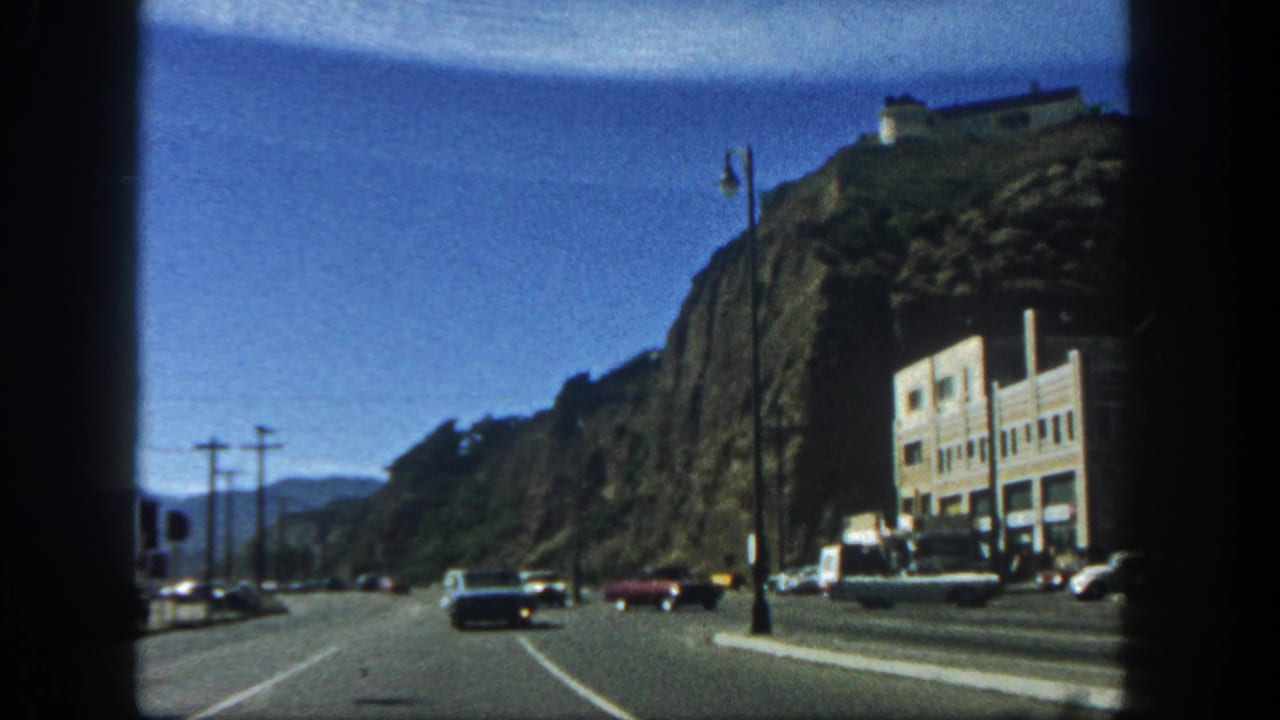 1962 San Diego road view with cars passing a rocky hillside, utility poles, streetlamps, and a pale building near the pavement.