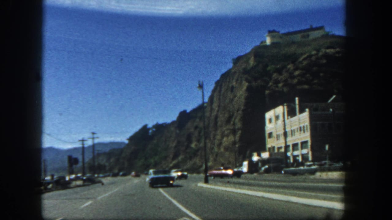 A road curves past a cliffside with cars, poles, and a large roadside building.