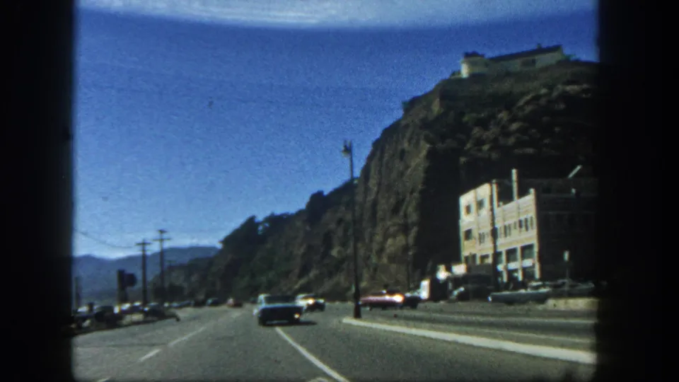 1962 San Diego driving view past a cliffside, with cars on the roadway and a large pale building at the base of the rock face.