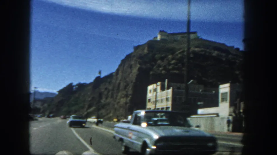 1962 San Diego roadside view with a classic car in the foreground, a rocky hill above, and buildings along the edge of the road.