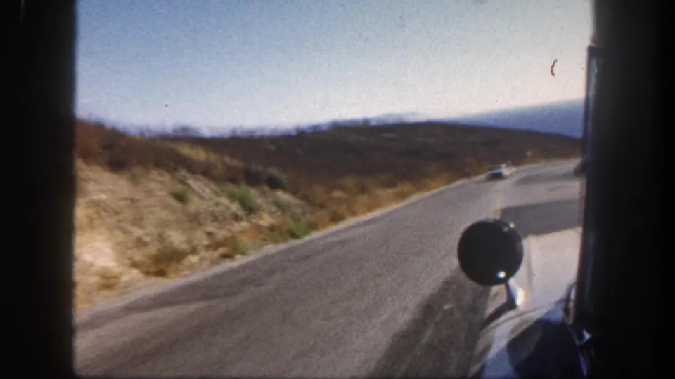 Passenger-side 8mm view from a moving vehicle on a sunlit road, with dry hills, pale sky, and the car's round side mirror visible.