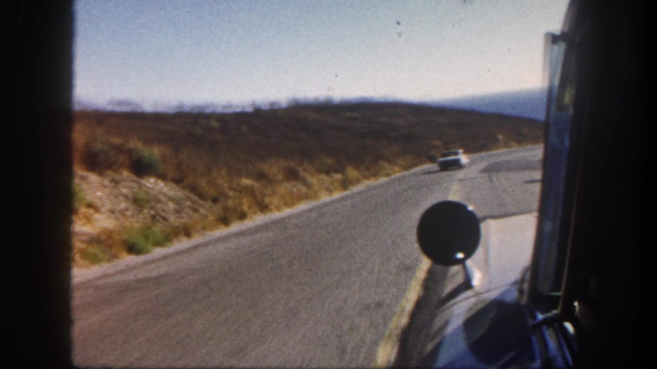 1962 San Diego-area 8mm view from a moving car on a winding road, with dry roadside brush, distant blue water, and a small car ahead.