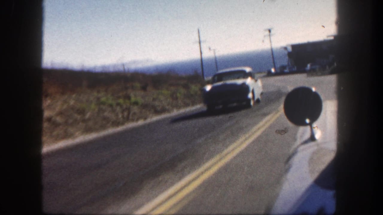 Oncoming white car passes on a two-lane road beside utility poles and dry open land.