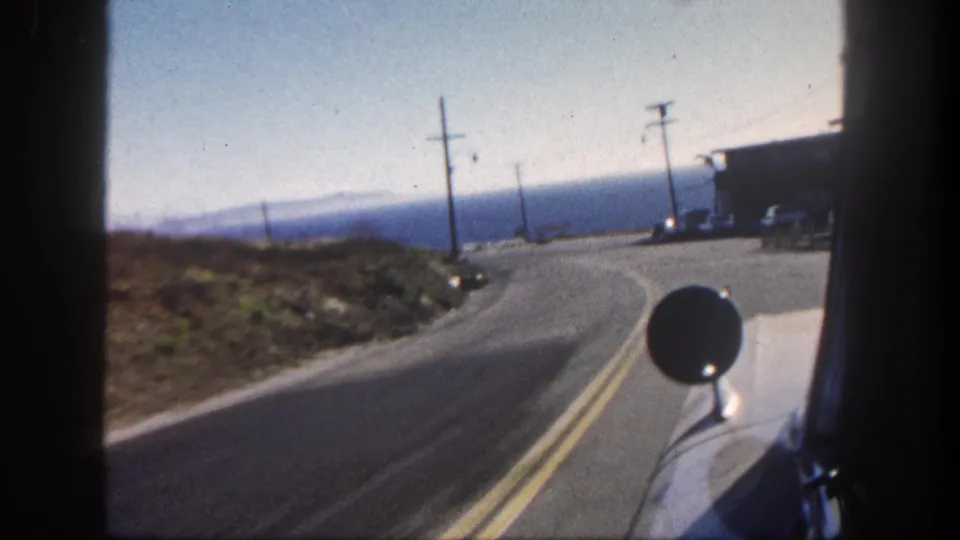 1962 San Diego-area 8mm frame showing a curving two-lane road, utility poles, dry slope, roadside structures, and blue water beyond.
