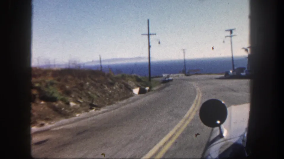 1962 San Diego-area car-window view of a downhill curve, utility poles, dry roadside banks, distant water, and part of the filming vehicle.