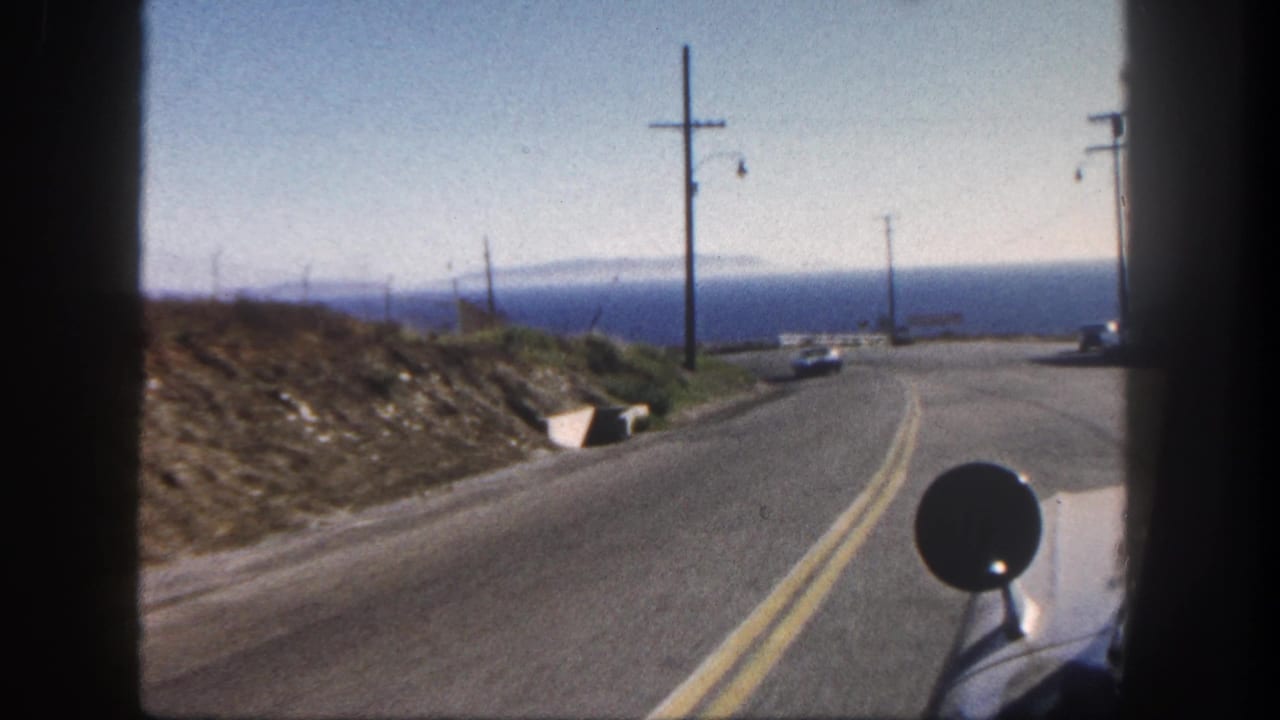 1962 San Diego coastal road seen from a vehicle, with double yellow lines, utility poles, dry hillside, and ocean beyond.