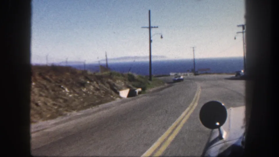 1962 San Diego coastal road seen from a vehicle, with double yellow lines, utility poles, dry hillside, and ocean beyond.