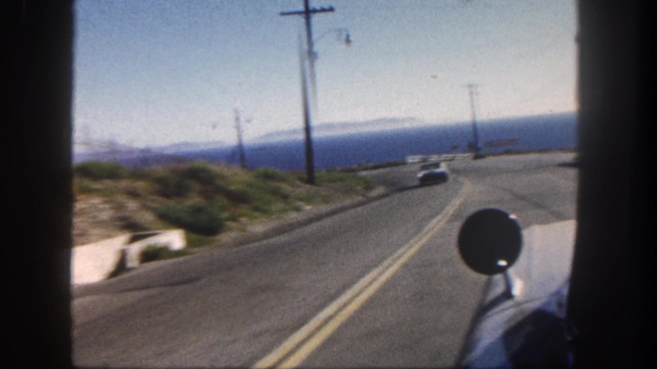 Vehicle POV along a 1962 San Diego coastal road, showing a bend, roadside poles, ocean horizon, and a white car ahead.