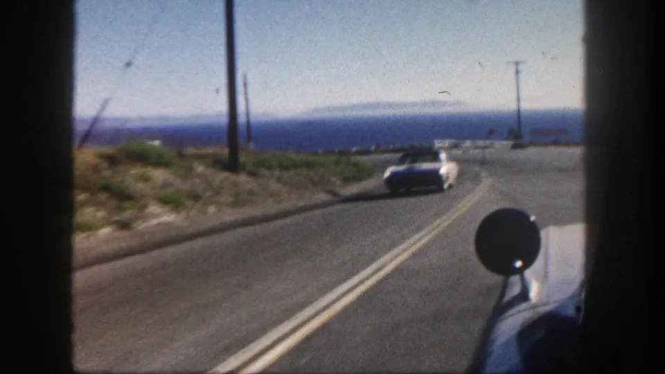 1962 San Diego vehicle POV showing an oncoming car on a coastal two-lane road with ocean, poles, and dry roadside vegetation.