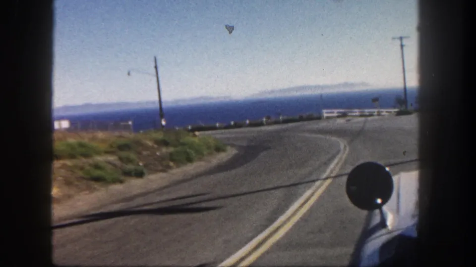 1962 San Diego coastal driving POV with a curving road, Pacific Ocean horizon, roadside rail, utility poles, and part of the car mirror visible.