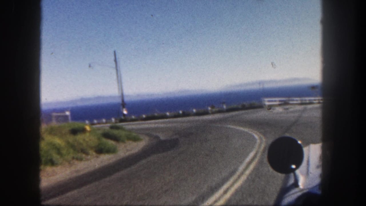 San Diego coastal highway POV from 1962 showing the road curving toward a white rail with the Pacific Ocean and car mirror in view.