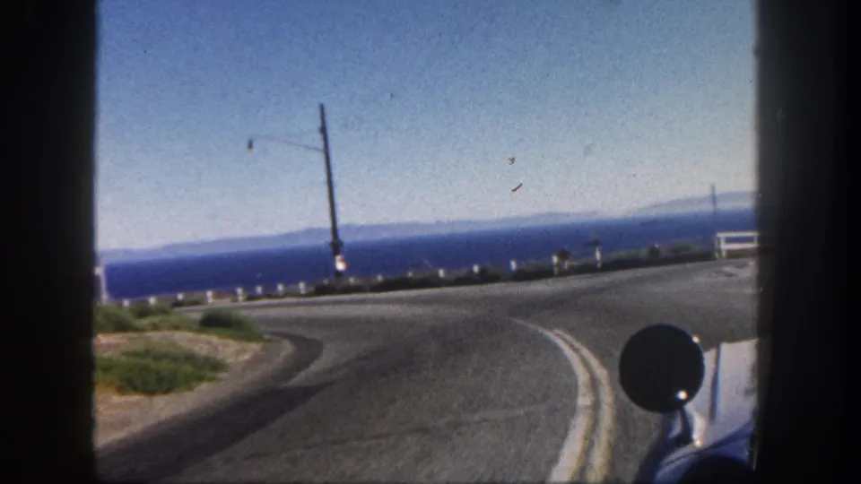 1962 home-movie driving view in San Diego with a coastal road, ocean horizon, roadside posts, utility pole, low vegetation, and vehicle mirror.