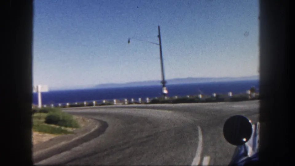 Late-frame 1962 San Diego coastal road POV showing a bend in the highway, ocean backdrop, roadside posts, utility pole, and the car mirror.