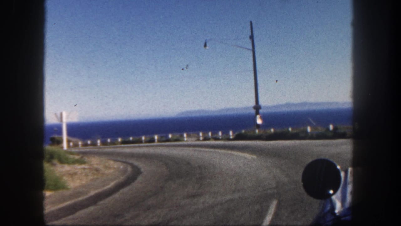 Curving coastal road seen from a moving vehicle with ocean, guardrail, utility poles, and vehicle mirror.