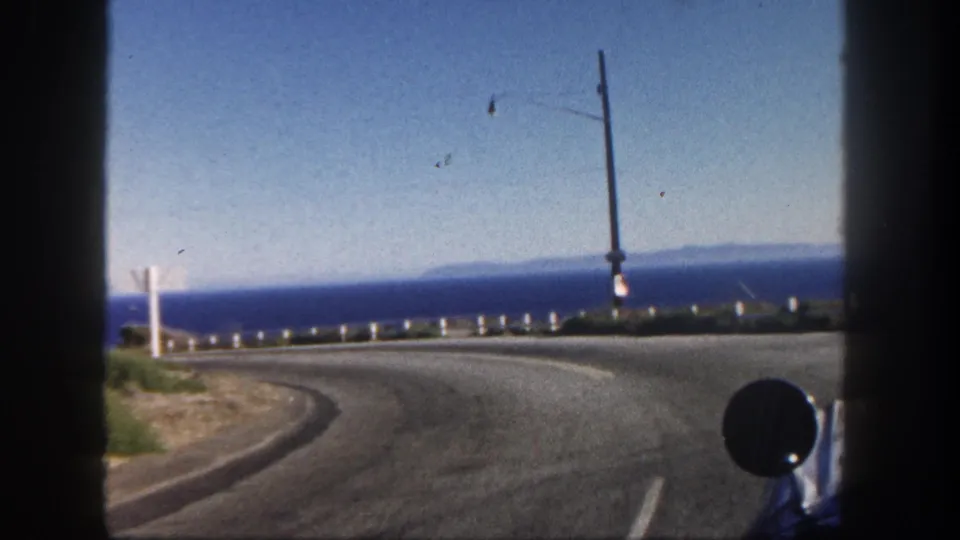 1962 San Diego coastal drive view showing a curving asphalt road, blue ocean horizon, white guardrail, utility poles, and a vehicle mirror.